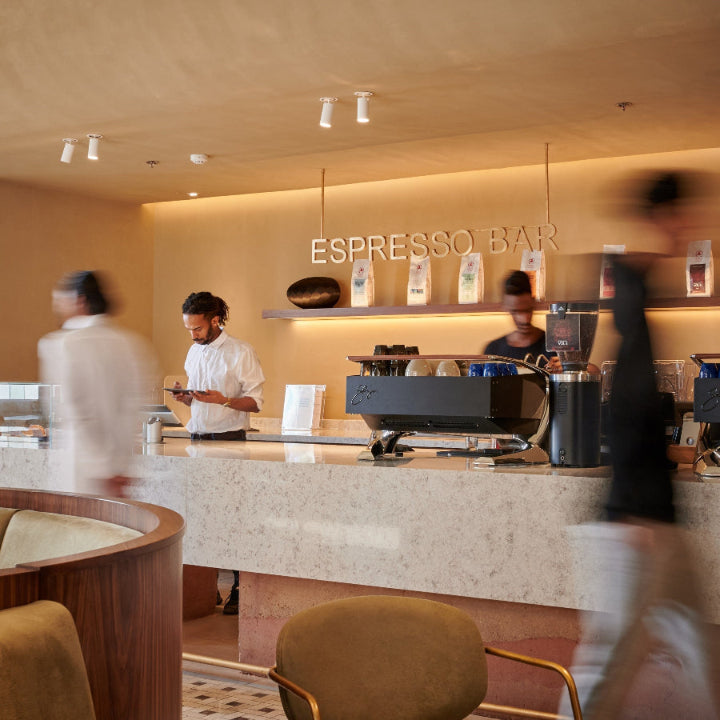 An elegant marble coffee bar with an espresso machine and a barista working to prepare coffee. and wall mounted Shelves holding coffee beans and mugs. And customers passing.