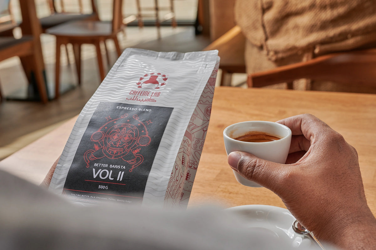 a coffee drinker holding a coffee bean bag of the better barista on the left hand and an espresso made by the same beans in the right hand and he is sitting on one of the tables of Caffeine Lab Café