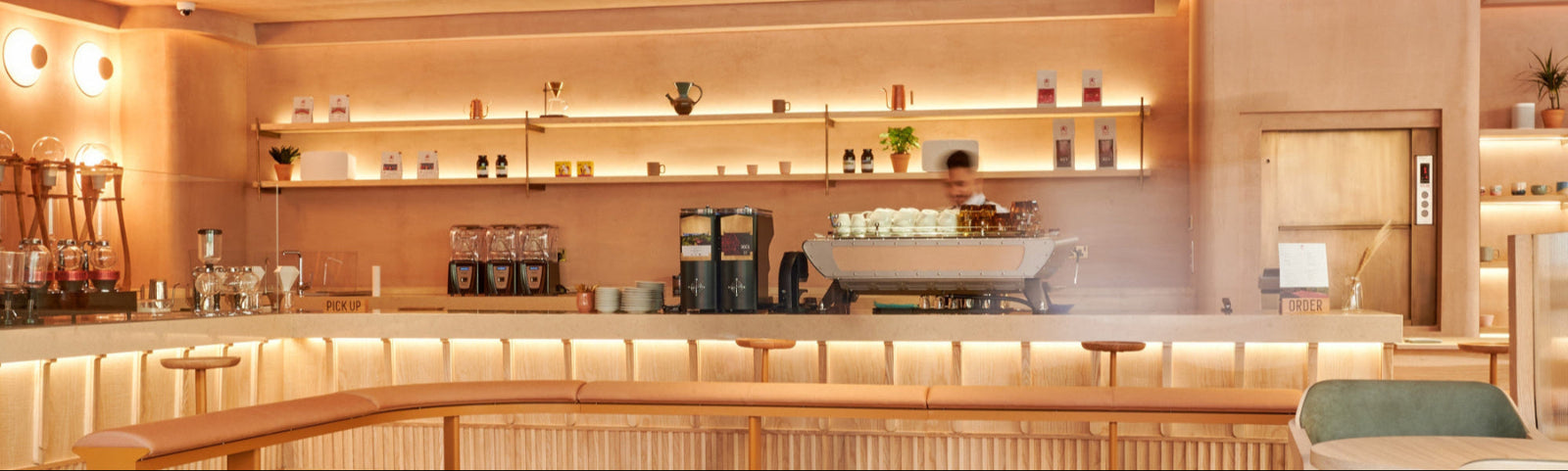An elegant sandy coffee bar with an espresso machine and a barista working to prepare coffee. and wall mounted Shelves holding coffee beans and mugs